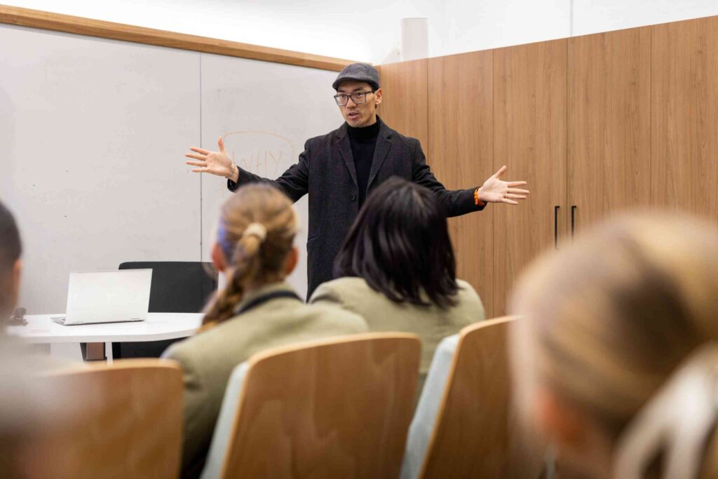 Person in a black outfit giving a lecture to seated students in a classroom.